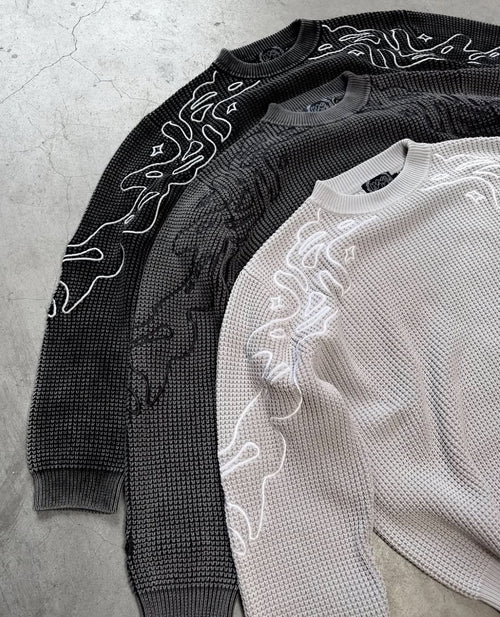 Three sweaters, one black and one gray, with intricate white patterns on a concrete surface.