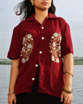 Person wearing a maroon shirt with floral embroidery by a body of water