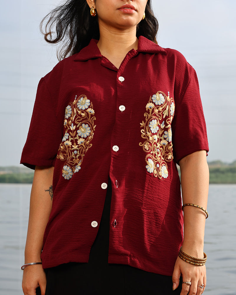 Person wearing a maroon shirt with floral embroidery by a body of water
