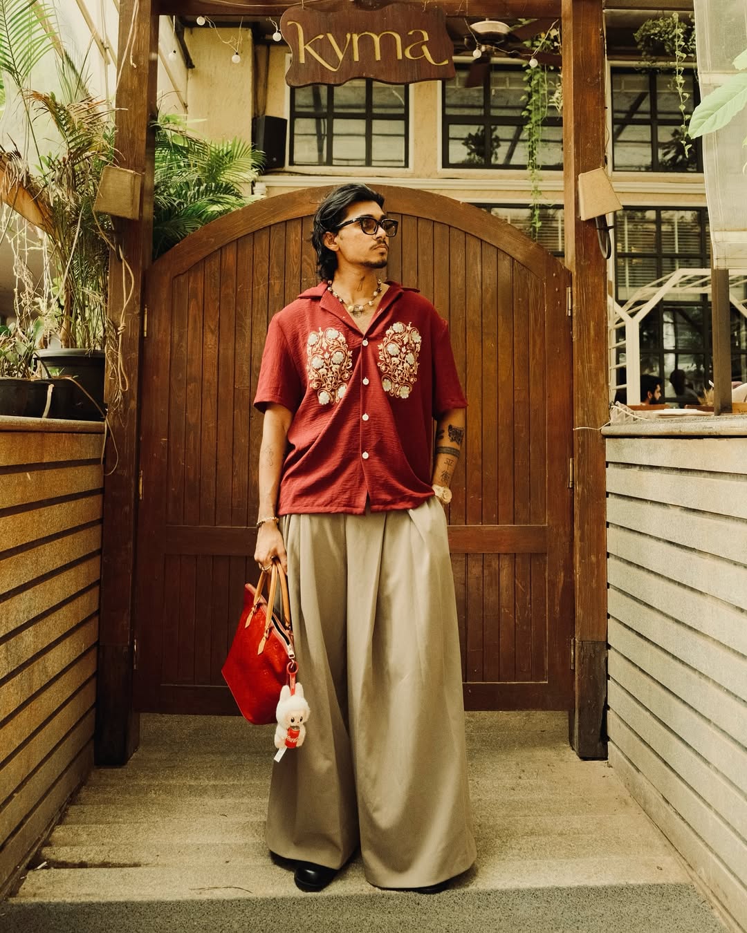 Person wearing a red shirt with floral patterns and beige pants, standing in front of a wooden door with 'kyma' branding.