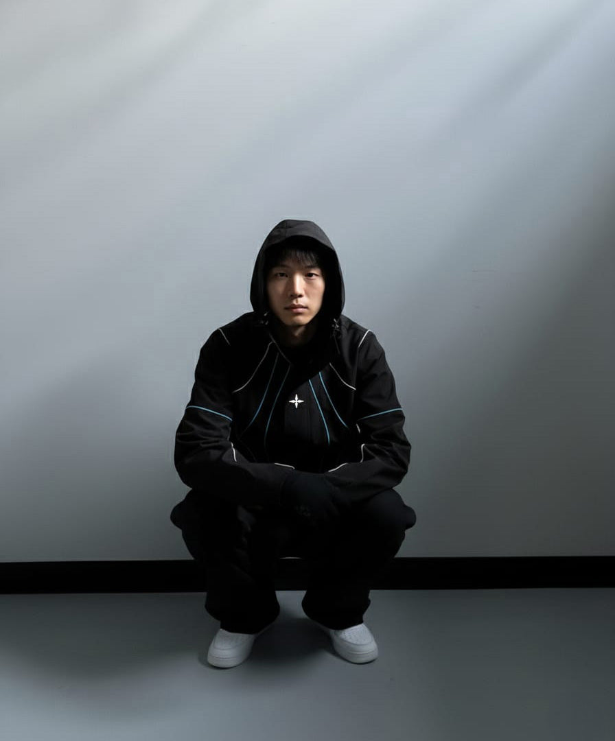Person wearing a black hoodie and pants sitting on a gray floor against a white wall.
