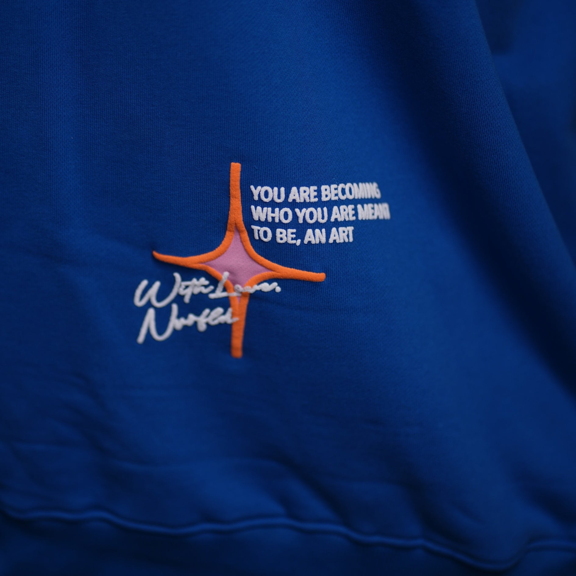 Close-up of NVRFLD blue graphic hoodie with orange and white embroidered inspirational text design