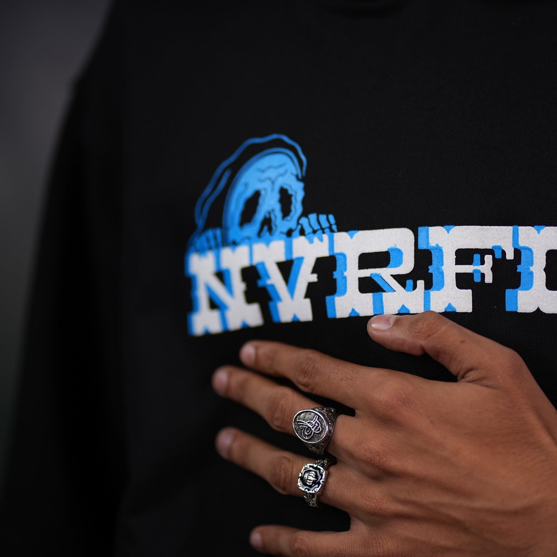 Close-up of NVRFLD black edgy streetwear hoodies with blue skull logo and hand wearing silver rings